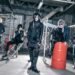 The photo shows a group of five people in black clothing standing inside what seems to be an industrial space. All five musicians appear to be looking off into different directions while maintaining their composure despite being surrounded by such an industrial setting.