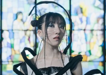 A close up photo of Haru Nemuri in front of a stained-glass window. Haru Nemuri has her hair in two knots and a couple of small braids. She is wearing artistic clothing with the colours black and white.  Photography by Maria Nanashima.