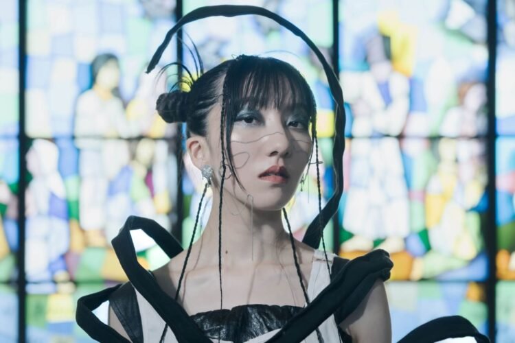 A close up photo of Haru Nemuri in front of a stained-glass window. Haru Nemuri has her hair in two knots and a couple of small braids. She is wearing artistic clothing with the colours black and white.  Photography by Maria Nanashima.