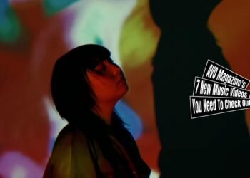 Still from OTO MAYUMI's music video for the song "だって (Datte)". The singer-songwriter is standing in front of a wall on which a beamer shines seemingly psychedelic-like patterns. As she stands between the beamer and wall, a shadow of herself is created on the wall. She looks up after dancing. The video is directed by Pierluigi Patella.