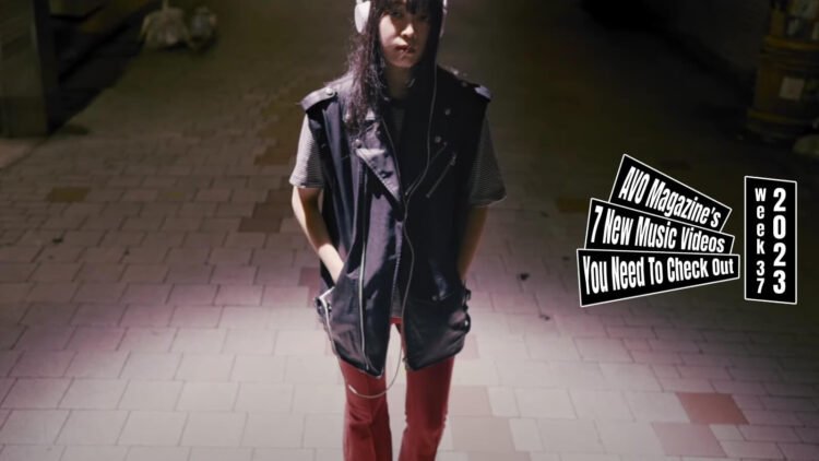 Still from the music video "ティーネイジャーの夜に (Teenager no Yoru Ni)" by 変心芙蓉 (Henshin Fuyou). The vocalist of Henshin Fuyou walks around in a street in the middle of the night, she wears dark-coloured jacket and red pants and earphones. A street light is shining light on her while she is having her hands in her pockets and looks toward the camera.