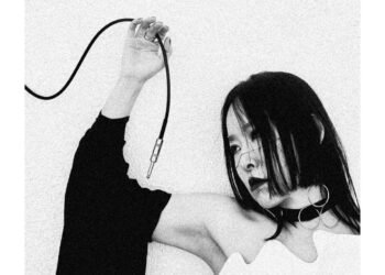 A monochrome photo of Yuko Araki, dressed in white and black, while she is posing and holding a cable that is part of the electronic gear that she uses to make her music. The background behind her is plain white, allowing for all attention to remain on the subject in focus. Photography by Room40/Lawrence English