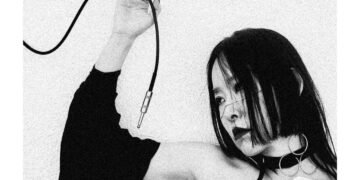 A monochrome photo of Yuko Araki, dressed in white and black, while she is posing and holding a cable that is part of the electronic gear that she uses to make her music. The background behind her is plain white, allowing for all attention to remain on the subject in focus. Photography by Room40/Lawrence English