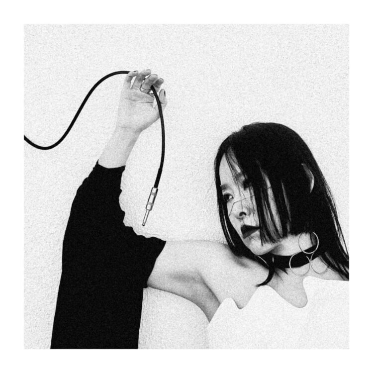 A monochrome photo of Yuko Araki, dressed in white and black, while she is posing and holding a cable that is part of the electronic gear that she uses to make her music. The background behind her is plain white, allowing for all attention to remain on the subject in focus. Photography by Room40/Lawrence English