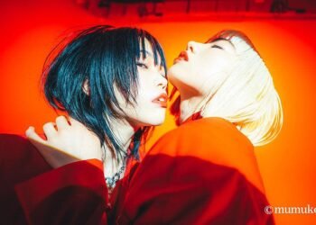 The musician AiNA THE END and dancer AOI YAMADA are dressed in what seems orange/red-coloured clothing and are standing very near to each other, almost in a hugging position. While AOI YAMADA has her eyes closed and her head up, while AiNA THE END eyes are looking towards the camera but her head is not facing the camera. Photography by mumuko