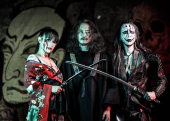 A couple of the members of SIGH (in total there are three people in the photo), including founder Mirai Kawashima (dressed in black) and Dr. Mikannibal (dressed in traditional clothing but edited it in wearing it off the shoulders and with a bloody face), standing next to each other, two of the are holding swords while all are looking into the direction of the photographer. Behind them is a big tapestry or mural with the album cover of "Shiki".