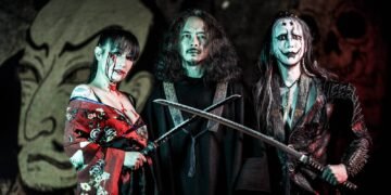 A couple of the members of SIGH (in total there are three people in the photo), including founder Mirai Kawashima (dressed in black) and Dr. Mikannibal (dressed in traditional clothing but edited it in wearing it off the shoulders and with a bloody face), standing next to each other, two of the are holding swords while all are looking into the direction of the photographer. Behind them is a big tapestry or mural with the album cover of "Shiki".