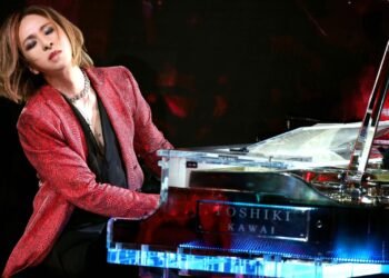 This photo captures an intimate moment between the musician YOSHIKI and his Kawai piano on stage. He wears a red jacket and a black blouse. YOSHIKI has his eyes closed and appears to be deeply focused on his music.