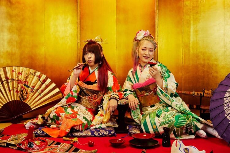 The picture shows the two women of AINAKANNA sitting on the ground and surrounded by traditional decorations (fan, umbrella, swords, masks, toys, tableware) and a gold-coloured wall. They are dressed in Japanese traditional clothing, kimono, and one is holding a pipe, while the other holds a small cup.