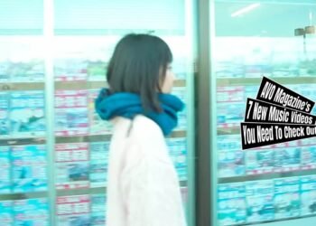 Still from DaimonOrchestra's music video for "キシロカイン (Xylocaine)". One of the members of DaimonOrchestra is walking in the streets past shops with windows plastered with advertisements. She wears a white wool jumper and a blue scarf. The still is a bit blurry because the camera and the musician are both moving.