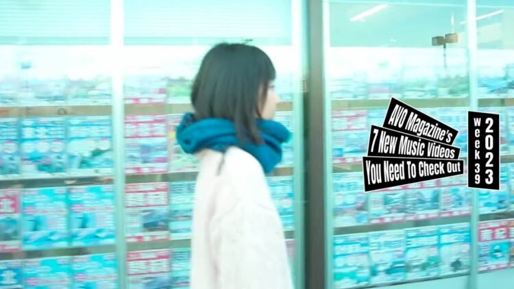Still from DaimonOrchestra's music video for "キシロカイン (Xylocaine)". One of the members of DaimonOrchestra is walking in the streets past shops with windows plastered with advertisements. She wears a white wool jumper and a blue scarf. The still is a bit blurry because the camera and the musician are both moving.