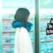Still from DaimonOrchestra's music video for "キシロカイン (Xylocaine)". One of the members of DaimonOrchestra is walking in the streets past shops with windows plastered with advertisements. She wears a white wool jumper and a blue scarf. The still is a bit blurry because the camera and the musician are both moving.