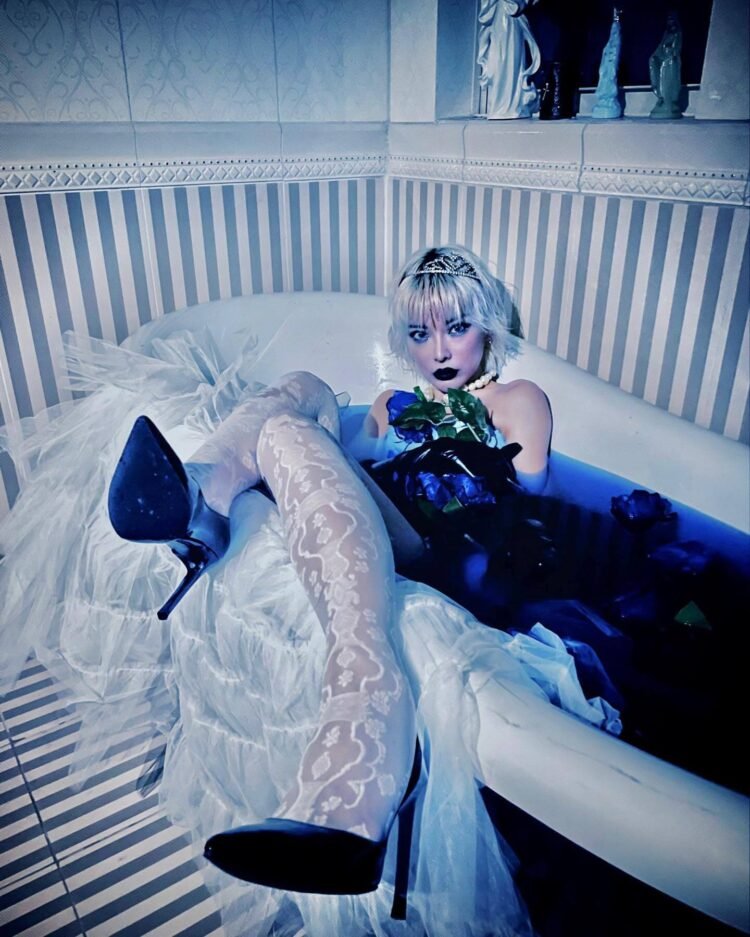 Nana Kitade is dressed in a white dress and high heels and sits in a bathtub filled with blue water.
