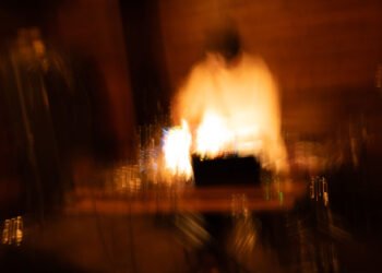 Artistic and blurry, out of focus photo of Phew in action (during a performance). The dominant colors are brown and black, while a white accent colour adds a subtle hint of vibrancy to this otherwise dark setting. The photo is taken by Masayuki Shioda.