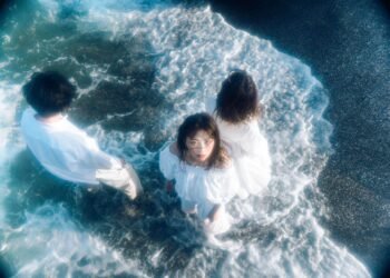 Three members of Tonari no Hanako are standing in the water, enjoying a sunny day. They are wearing white clothing. One of the members, is looking into the camera, while the others have their backs turned to the camera.