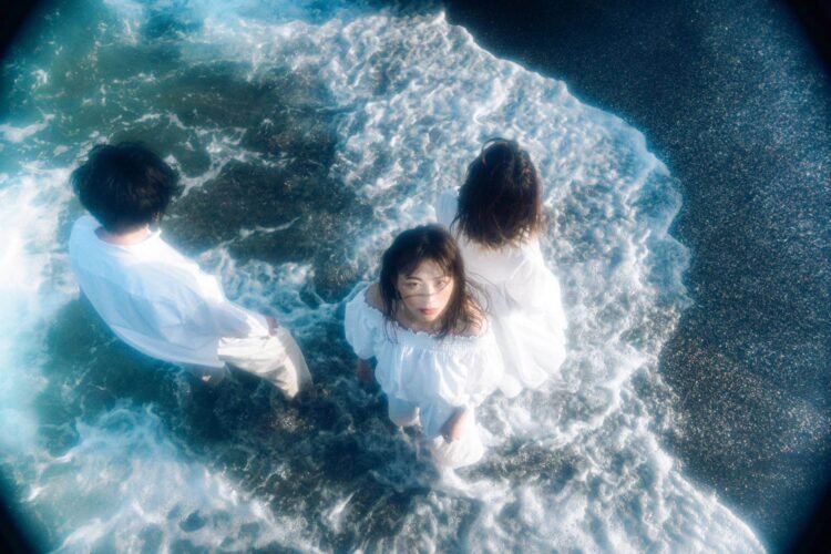 Three members of Tonari no Hanako are standing in the water, enjoying a sunny day. They are wearing white clothing. One of the members, is looking into the camera, while the others have their backs turned to the camera.