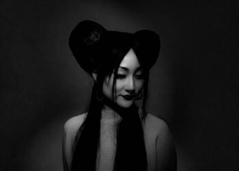 A monochrome close-up of Hinako Omori with two big buns in her hair but also has part of her hair loose. She wears a striped shirt and does not look into the camera but looks sideways down with a soft smile.