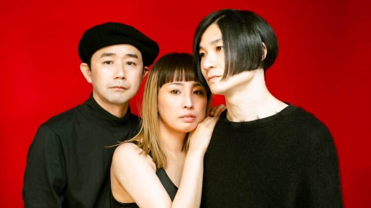 Group photo of the three members of MASS OF THE FERMENTING DREGS, standing, they are standing closely side by side.. All musicians are dressed in dark clothing and on the background is a red wall.