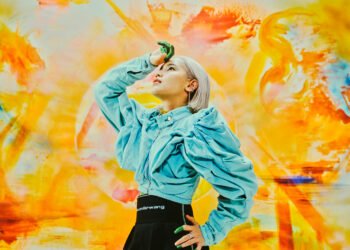 Colourful photo of singer-songwriter Anly posing in front of a painting by Karin Okoso. The painting shows a lot of yellow and orange hues while the musician dresses a blue denim blouse and black skirt. Anly's right hand is resting on her head while she looks up. Her fingertips are painted green and might give an idea that she has been working on a painting before taking the photo. Photo by Seiya Fujii.