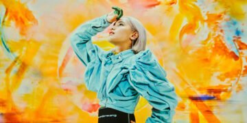 Colourful photo of singer-songwriter Anly posing in front of a painting by Karin Okoso. The painting shows a lot of yellow and orange hues while the musician dresses a blue denim blouse and black skirt. Anly's right hand is resting on her head while she looks up. Her fingertips are painted green and might give an idea that she has been working on a painting before taking the photo. Photo by Seiya Fujii.