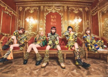 The image shows the idol You’ll Melt More! sitting on a a red with golden accents couch or seat. in a room. Each group member are dressed in clothing that features predominantly yellow, white and black accents and their own member colour. In the background, elaborate gold-trimmed patterns against a red-coloured wall contributes to the luxurious ambiance.