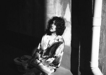 A black and white photograph of the rock musician DURAN seated on the floor against a textured wall. The musician's attire consists of a tie-dyed shirt and he is holding a guitar by his side. The specific details of DURAN's expression are obscured by shadows and his curly hair.