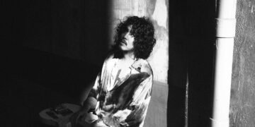 A black and white photograph of the rock musician DURAN seated on the floor against a textured wall. The musician's attire consists of a tie-dyed shirt and he is holding a guitar by his side. The specific details of DURAN's expression are obscured by shadows and his curly hair.