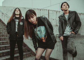 This photo displays three members of a metal band called GUNGIRE, standing in front of a grey wall near a flight of stairs. They are all dressed in black attire—two men in the back and one woman in front.