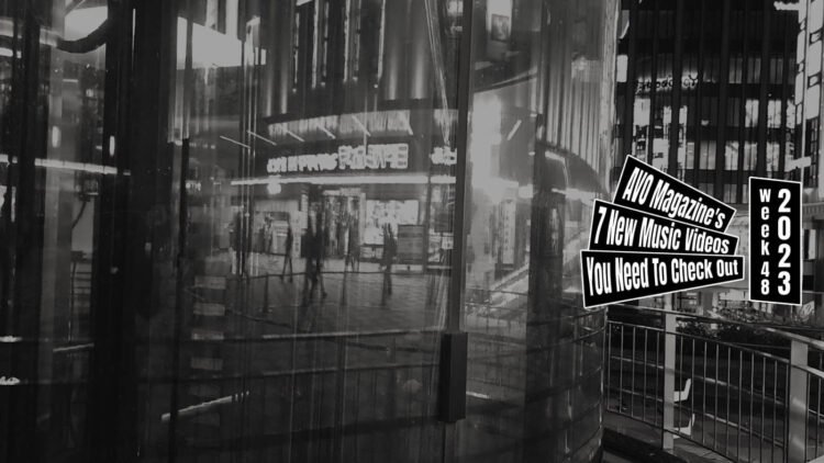 A still from the band CULTE's music video for the song "Sugarmuffin". A cityscape in black and white with a focus on a building or wall of glass and the reflection of neon lights from shops behind the person filming this and the people walking past.