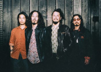 The photo features the four men of krautrock band Minami Deutsch. They are standing in close proximity to each other against a wooden backdrop, dressed casually, some are wearing leather jackets.