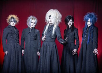 The photo depicts five members of the Japanese visual kei metal band Zeke Deux, positioned in front of a red curtain backdrop. Each member is uniquely dressed in black attire - seemingly a long jacket and under it a button-down shirt and tie - with diverse hair colours and hairstyles. They are all standing in a line, with vocalist Kakeru standing a bit more in front while holding his arm out showing his hand jewellery with what seems metal nails.