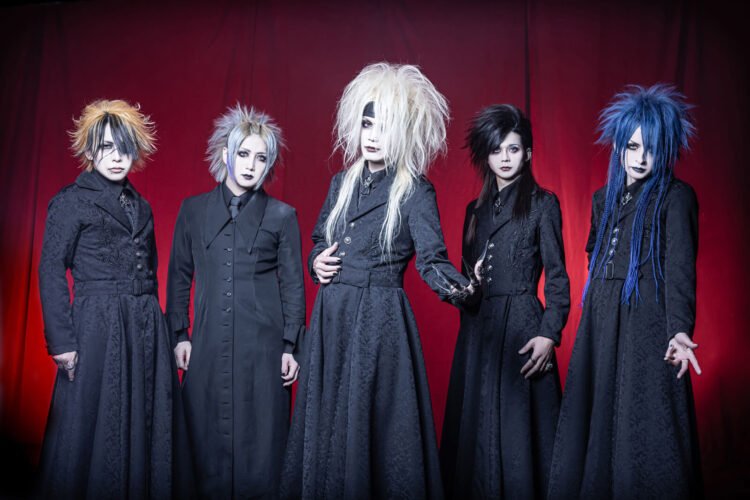 The photo depicts five members of the Japanese visual kei metal band Zeke Deux, positioned in front of a red curtain backdrop. Each member is uniquely dressed in black attire - seemingly a long jacket and under it a button-down shirt and tie - with diverse hair colours and hairstyles. They are all standing in a line, with vocalist Kakeru standing a bit more in front while holding his arm out showing his hand jewellery with what seems metal nails.