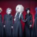 The photo depicts five members of the Japanese visual kei metal band Zeke Deux, positioned in front of a red curtain backdrop. Each member is uniquely dressed in black attire - seemingly a long jacket and under it a button-down shirt and tie - with diverse hair colours and hairstyles. They are all standing in a line, with vocalist Kakeru standing a bit more in front while holding his arm out showing his hand jewellery with what seems metal nails.