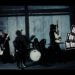Still from the music video "アリガトウ (Arigatou)" by the band Luin. The still depicts the band Luin performing in a space with dim lighting making the atmosphere moody. The musicians, dressed in black, are playing various instruments including a bass guitar, the drums, and guitars.