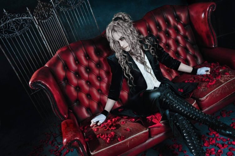 KAMIJO is seated on a red leather sofa surrounded by red rose petals that are also scattered under and around the sofa. He is clad in a white shirt with a black velvet jacket, black and white-coloured gloves, knee-length black boots, and is looking at the camera.