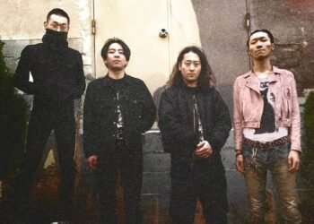 The four band members of KLONNS stand side by side in front of a concrete wall with a door. The wall seems to be painted in greyish and off-white colours with a bit of red. Three of the musicians are dressed entirely in black clothing while the fourth band member wears jeans and a light pink coloured jacket over a white shirt with a black cat on it. Some filters have been applied to the photo which clearly shows some noise in the picture and also makes it look a bit dated. Photo by Chihiro Yoshikawa.