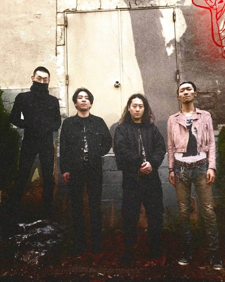 The four band members of KLONNS stand side by side in front of a concrete wall with a door. The wall seems to be painted in greyish and off-white colours with a bit of red. Three of the musicians are dressed entirely in black clothing while the fourth band member wears jeans and a light pink coloured jacket over a white shirt with a black cat on it. Some filters have been applied to the photo which clearly shows some noise in the picture and also makes it look a bit dated. Photo by Chihiro Yoshikawa.