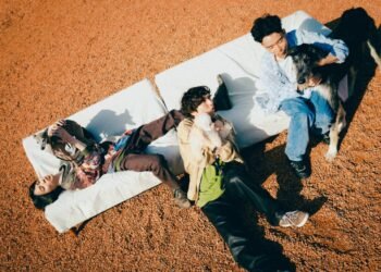 The three musicians of Helsinki Lambda Club lying and sitting on white retangle pillows that are placed on a sandy location. They are closely huddled together and holding dogs.

L-R Taiki Kumagai (guitar), Kaoru Hashimoto (vocals/guitar), Kodai Inaba (bass)

Photo by: Renzo Masuda