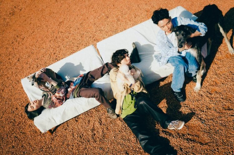 The three musicians of Helsinki Lambda Club lying and sitting on white retangle pillows that are placed on a sandy location. They are closely huddled together and holding dogs.

L-R Taiki Kumagai (guitar), Kaoru Hashimoto (vocals/guitar), Kodai Inaba (bass)

Photo by: Renzo Masuda