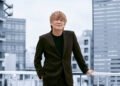 Hironobu Kageyama dressed in a black suit jacket and sweater is standing on what seems to be a rooftop, with one of his hands resting on a white railing. Behind him, there's a cityscape with multiple buildings. © HIGHWAY STAR Inc.