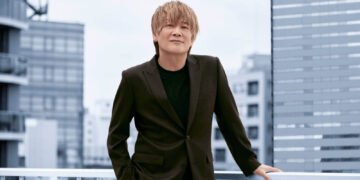 Hironobu Kageyama dressed in a black suit jacket and sweater is standing on what seems to be a rooftop, with one of his hands resting on a white railing. Behind him, there's a cityscape with multiple buildings. © HIGHWAY STAR Inc.