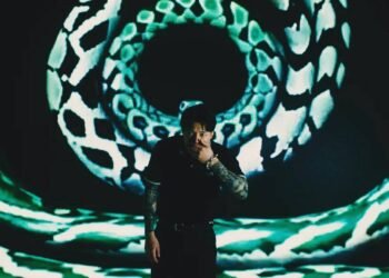 Ryo Kinoshita stands in front of a screen that shows what seems to be snakeskin patterns presented in circles. He wears a black coloured shirt and pants and is holding his left hand in front of his mouth.