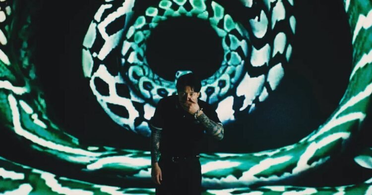 Ryo Kinoshita stands in front of a screen that shows what seems to be snakeskin patterns presented in circles. He wears a black coloured shirt and pants and is holding his left hand in front of his mouth.