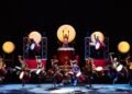 The drummers of Yamato are standing on a dark stage during a performance performing on their taiko drums, some are performing on a heightened stage, while others are performing in the front. Photo by © Hiroshi Seo | provided by BB Promotion