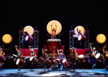 The drummers of Yamato are standing on a dark stage during a performance performing on their taiko drums, some are performing on a heightened stage, while others are performing in the front. Photo by © Hiroshi Seo | provided by BB Promotion