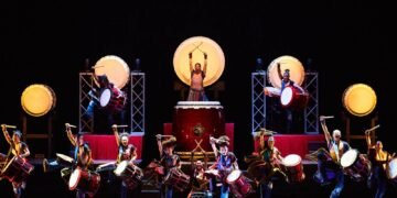 The drummers of Yamato are standing on a dark stage during a performance performing on their taiko drums, some are performing on a heightened stage, while others are performing in the front. Photo by © Hiroshi Seo | provided by BB Promotion