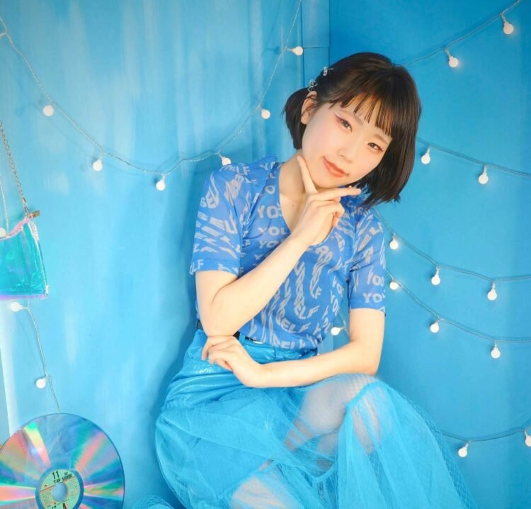 In this photo, Yukka is dressed in a blue outfit and seated in a blue-themed room, sitting on a blue bench with her right hand supporting her head while her other arm is resting on her left leg. Photo by Gentoku Ishii