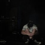The photo depicts a scene set in a dimly lit room, highlighting BGGB who is sitting cross-legged on the floor while his face is obscured by a black coloured balaclava.