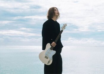 Ichika Nito dressed in black holds his electric guitar in his left hand while looking off to the side. The photograph is taken in such a way that there are only clouds visible behind the musician.