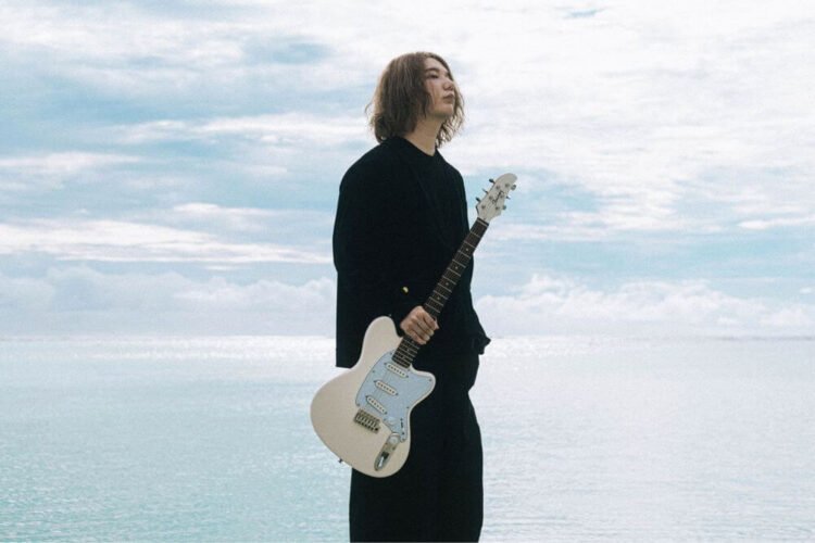 Ichika Nito dressed in black holds his electric guitar in his left hand while looking off to the side. The photograph is taken in such a way that there are only clouds visible behind the musician.